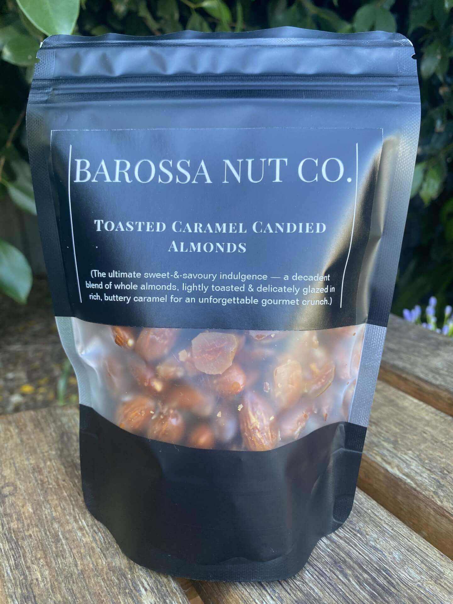 Bag of Barossa Nut Co. toasted caramel candied almonds on a wooden surface with greenery in the background.